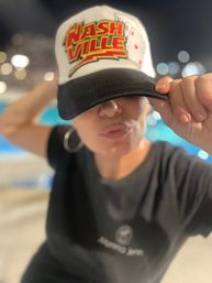 Playful person by a lit pool at night pulling down a white-and-black cap reading "Nashville," puckering lips, wearing a hoop earring and black t‑shirt with bokeh lights in the background.