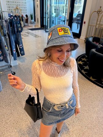 Smiling shopper in a white lace top and denim mini skirt with embellished belt, wearing a denim bucket hat with a colorful patch and carrying a black handbag inside a stylish fashion boutique with clothing racks and marble floor.