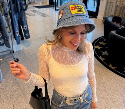 Smiling shopper in a white lace top and denim mini skirt with embellished belt, wearing a denim bucket hat with a colorful patch and carrying a black handbag inside a stylish fashion boutique with clothing racks and marble floor.