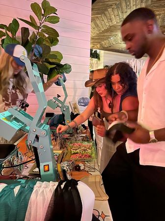 Young adults browsing jewelry, hats, and caps at an indoor pop-up market stall with teal heat-press machines, a potted fiddle-leaf plant, and warm wood-paneled ceiling lighting