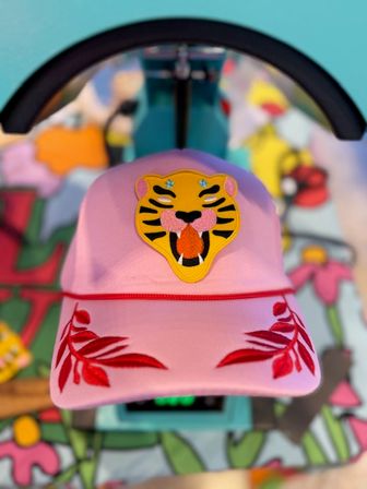 Close-up of a pink baseball cap with a bold embroidered yellow tiger face patch and red leaf embroidery on the brim, set against a colorful patterned background.