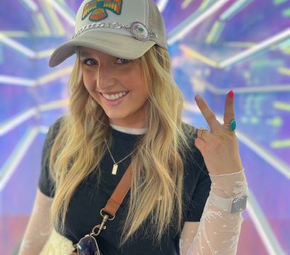 Smiling person in a trucker cap flashing a peace sign in a vibrant neon tunnel backdrop — black crop top, sheer long sleeves, crossbody bag and sunglasses, playful portrait.