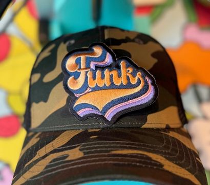 Camouflage baseball cap with a retro embroidered 'Funk' patch in orange and purple, placed on a turquoise hat press against a colorful blurred background.
