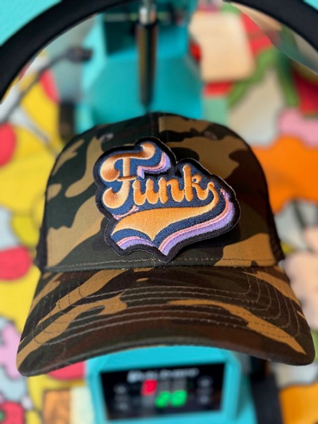 Camouflage baseball cap with a retro embroidered 'Funk' patch in orange and purple, placed on a turquoise hat press against a colorful blurred background.