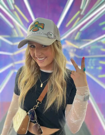 Smiling woman in a trucker hat and layered outfit flashes a peace sign in a vibrant purple neon/LED light tunnel, wearing a crossbody bag, sunglasses, and turquoise ring.