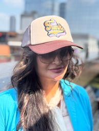 Sunlit urban portrait of a person wearing a retro 'Funk' trucker hat and sunglasses, long dark hair and a bright blue jacket with a blurred downtown skyline in the background.