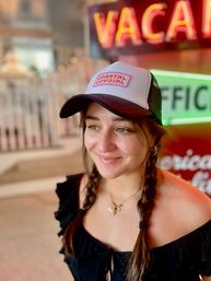 Portrait of a smiling woman with braided pigtails wearing a "Coastal Cowgirl" trucker hat and black off-shoulder top, with blurred neon signs in the background.