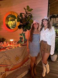 Two smiling women in casual western-chic outfits (baseball caps, cowboy boots) pose on a tropical-themed patio beside a snack table, fiddle-leaf plant, and round mirror under warm evening lights.