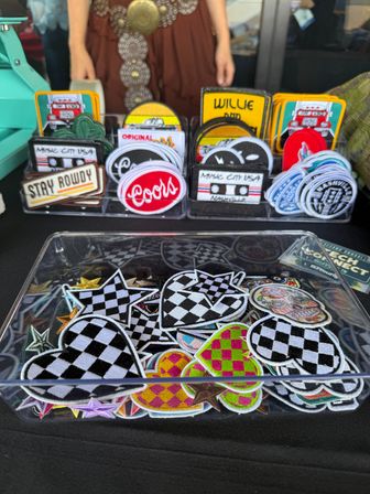 Colorful embroidered patches in clear trays on a market table — checkerboard hearts and stars, round retro-style and cassette tape designs at a Nashville music‑city vendor display.