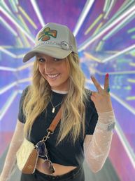 Smiling woman in a bird-embroidered trucker hat and black crop top layered over sheer lace sleeves, wearing a crossbody bag and sunglasses, flashing a peace sign in front of a colorful neon light tunnel backdrop.