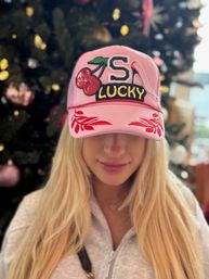 Blonde woman wearing a pink baseball cap with sequined cherry patch, large 'S' and 'LUCKY' lettering and red leaf accents, blurred Christmas tree and ornaments in the background