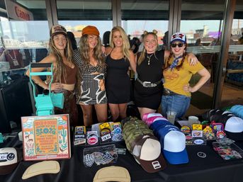 Five women pose behind a colorful vendor booth displaying trucker hats, patch designs and hat-making tools at a waterfront craft market