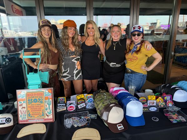 Five women pose behind a colorful vendor booth displaying trucker hats, patch designs and hat-making tools at a waterfront craft market