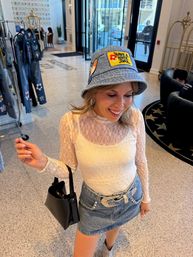 Smiling shopper in a boutique wearing a denim bucket hat with a 'Have a Nice Day' patch, sheer lace long-sleeve top, denim mini skirt with sparkly belt, carrying a black handbag near clothing racks and glass doors.