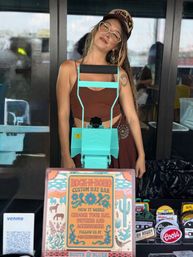 Smiling vendor in a cap and glasses using a turquoise hat-press at a boho custom-hat stall with colorful signage, patches, and a Venmo QR code at an outdoor market