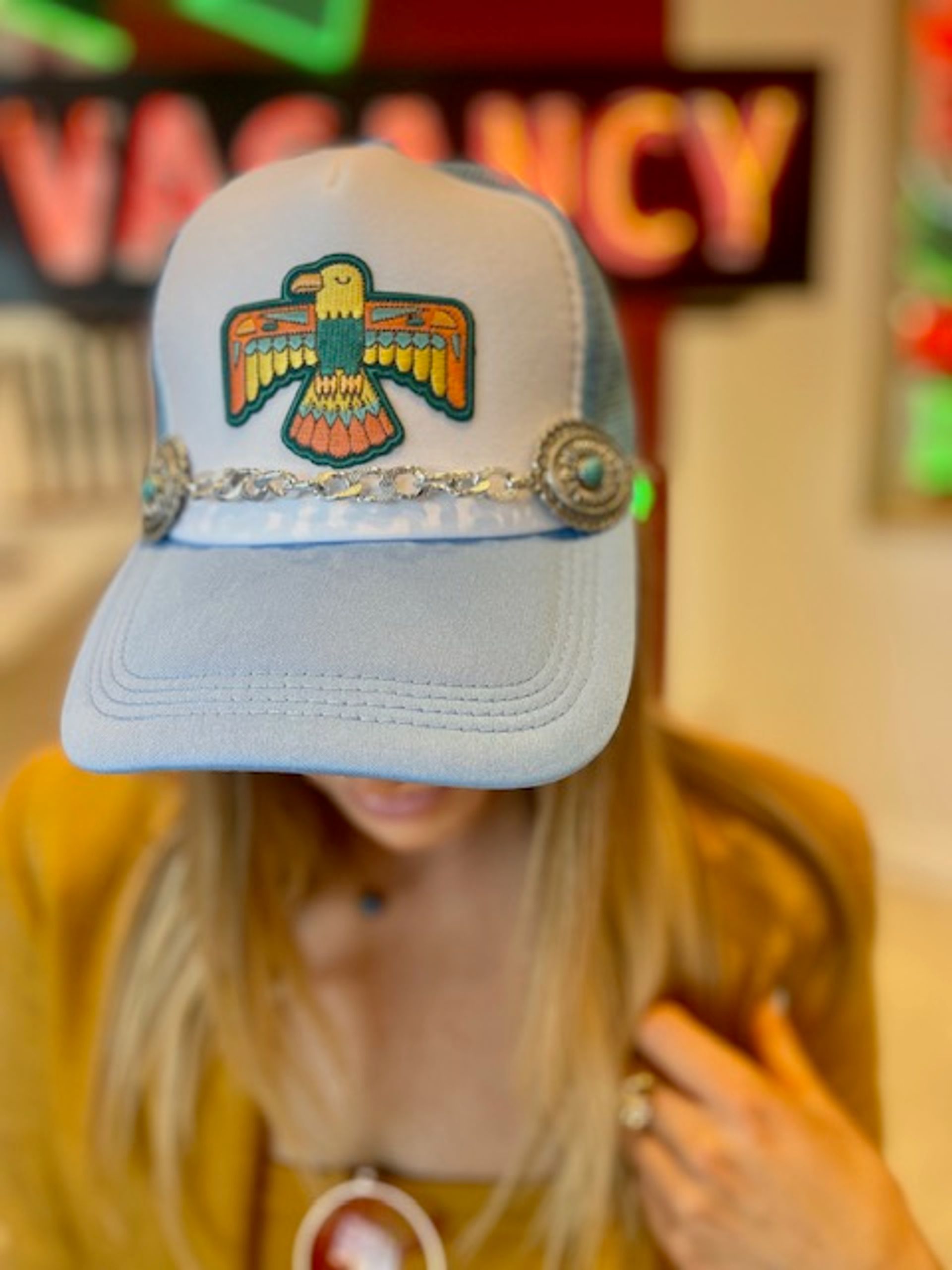 Close-up of a person wearing a light-blue trucker hat with a colorful embroidered eagle patch and silver concho trim, blonde hair visible, blurred neon "VACANCY" sign behind.