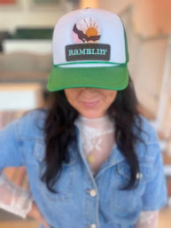 Casual indoor lifestyle portrait of a person in a denim jacket and lace top wearing a green-and-white trucker hat with RAMBLIN' sunset patch, blurred background