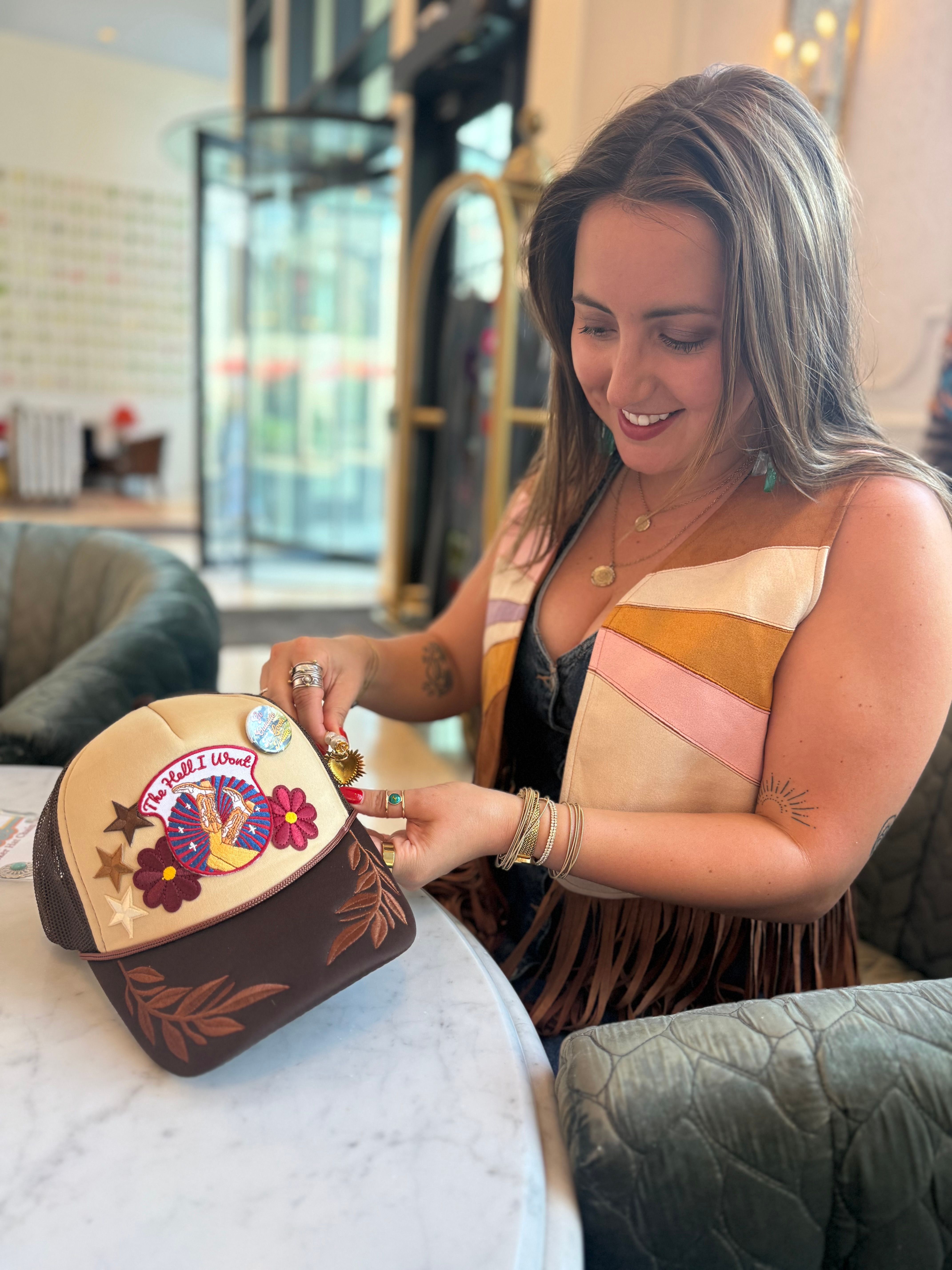 Smiling woman customizing a patch-covered trucker hat with pins and charms at a marble table in a bright hotel lobby