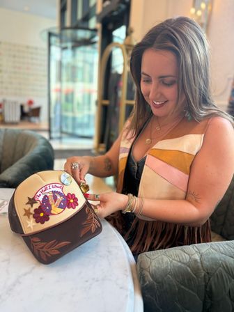 Smiling woman customizing a patch-covered trucker hat with pins and charms at a marble table in a bright hotel lobby