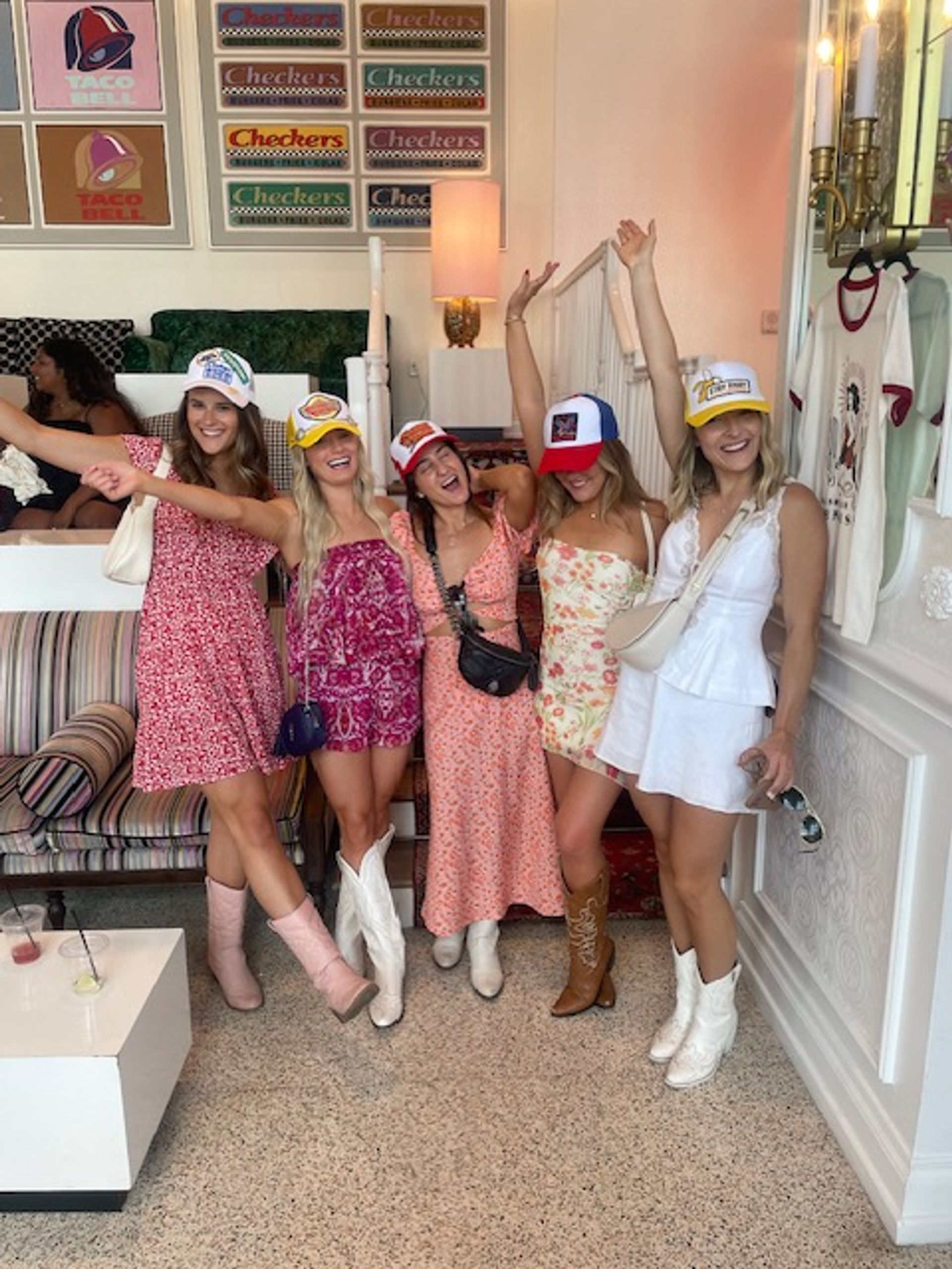 Five friends in colorful sundresses, cowboy boots and trucker hats smiling and posing with arms raised inside a bright boutique-style room with patterned sofa and wall art.