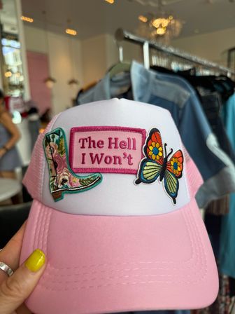 Pink-and-white trucker hat with embroidered cowboy boot and colorful butterfly patches and a pink patch reading "The Hell I Won't", held by a hand with yellow nail polish in a clothing boutique.