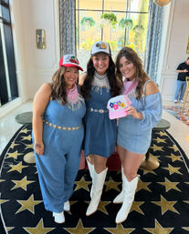 Three smiling women in denim outfits and bandanas wearing white cowboy boots and playful trucker hats, posing with a pink flower hat in a bright hotel lobby with a star-patterned rug and large windows.