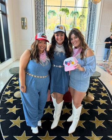 Three smiling women in coordinated denim outfits and white cowboy boots pose together indoors on a black star-pattern rug, wearing pink bandanas and colorful trucker hats in a bright hotel lobby with tall windows and hanging plants.