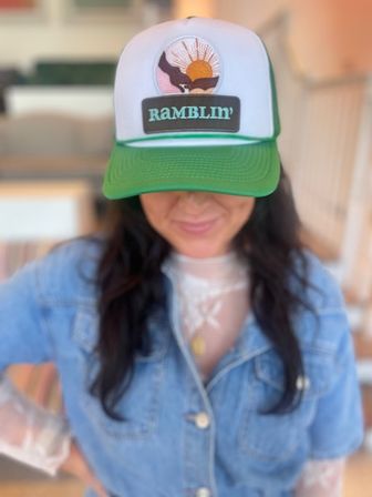 Person in a denim jacket wearing a green-and-white trucker hat with an embroidered patch that reads “Ramblin'” and shows a sunrise and bird, smiling down in a cozy indoor setting.