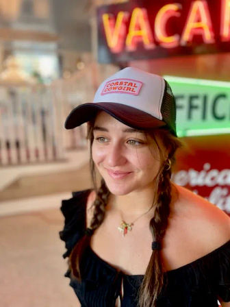 Smiling person with braided pigtails wearing a white-and-black trucker hat with a patch and an off-shoulder black top, seated in a neon-lit coastal diner with blurred vintage signs in the background.