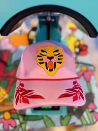 Pink embroidered baseball cap with a yellow roaring tiger patch and red leaf designs on the brim, photographed front-on against a colorful blurred mural background.