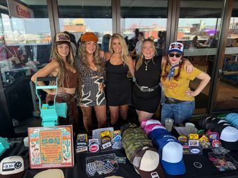 Five smiling women pose behind a vendor table of colorful trucker hats, caps, embroidered patches and stickers at a rooftop market, with a small hat press on the table and city buildings visible through glass behind them.