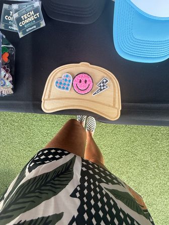 Overhead view of a vendor table on artificial turf with a wooden hat-shaped display holding three embroidered patches (blue-check heart, fuzzy pink smiley, black-and-white lightning bolt), a nearby blue cap and event stickers, and a person standing in a patterned skirt and checkerboard slip-on shoes.