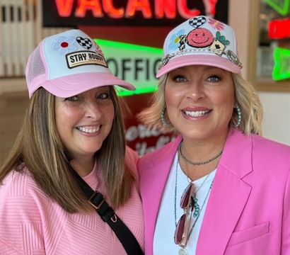 Two smiling women in pink trucker hats and pink outfits pose indoors with a neon "VACANCY" sign and retro motel-style decor in the background, bright cheerful portrait.