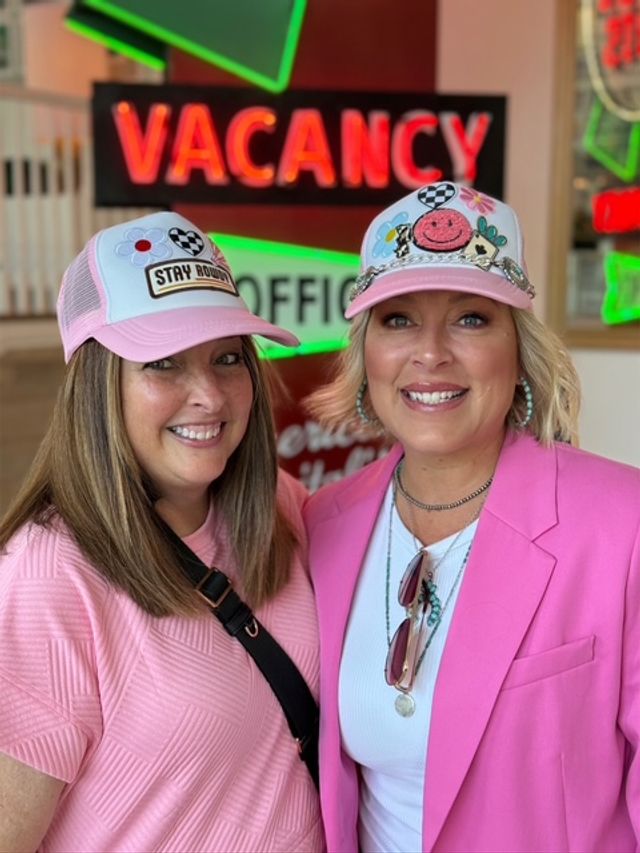 Two smiling women in pink trucker hats and pink outfits pose indoors with a neon "VACANCY" sign and retro motel-style decor in the background, bright cheerful portrait.