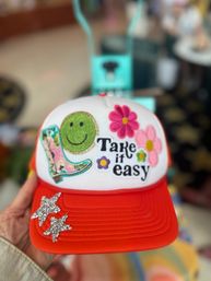 Bright red-and-white trucker hat held in hand with embroidered patches: a green smiley, pink flowers, patterned cowboy boot and 'Take it easy' text, plus two silver star embellishments on the brim against a blurred shop background