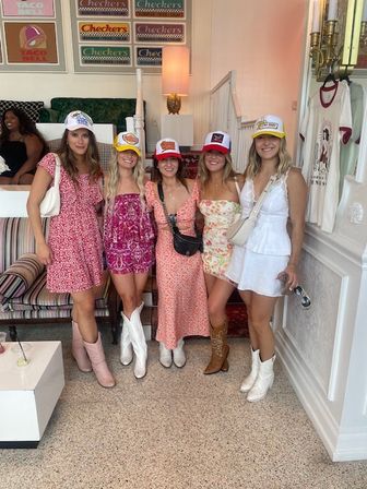 Five friends in floral summer dresses, trucker hats and cowboy boots posing indoors at a pastel, retro-themed boutique with framed vintage signs, a striped sofa and lamp.