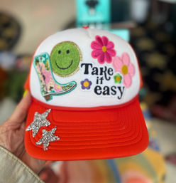 Bright red-and-white trucker hat held by a hand, decorated with a green smiley patch, pink flower patches, patterned cowboy boot patch, black "Take it easy" text patch, and two rhinestone star embellishments on the brim