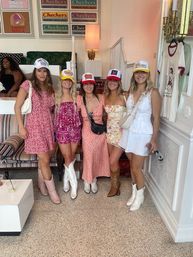 Group of five friends in summer dresses and trucker hats posing indoors in a retro-styled boutique café, wearing colorful cowboy boots and standing in front of vintage logo wall art