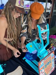 Two women leaning over a bright turquoise heat-press machine customizing a blue mesh trucker hat with patches and colorful pins at an outdoor craft vendor table.