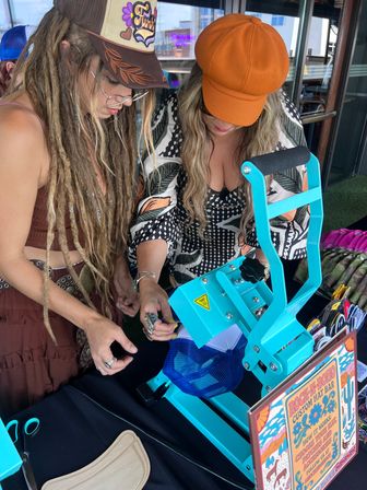 Two women leaning over a bright turquoise heat-press machine customizing a blue mesh trucker hat with patches and colorful pins at an outdoor craft vendor table.