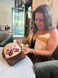 Smiling woman in a boho vest pins a sun-shaped enamel pin onto a brown-and-beige embroidered trucker hat decorated with flowers, stars and patches while seated at a marble café table.