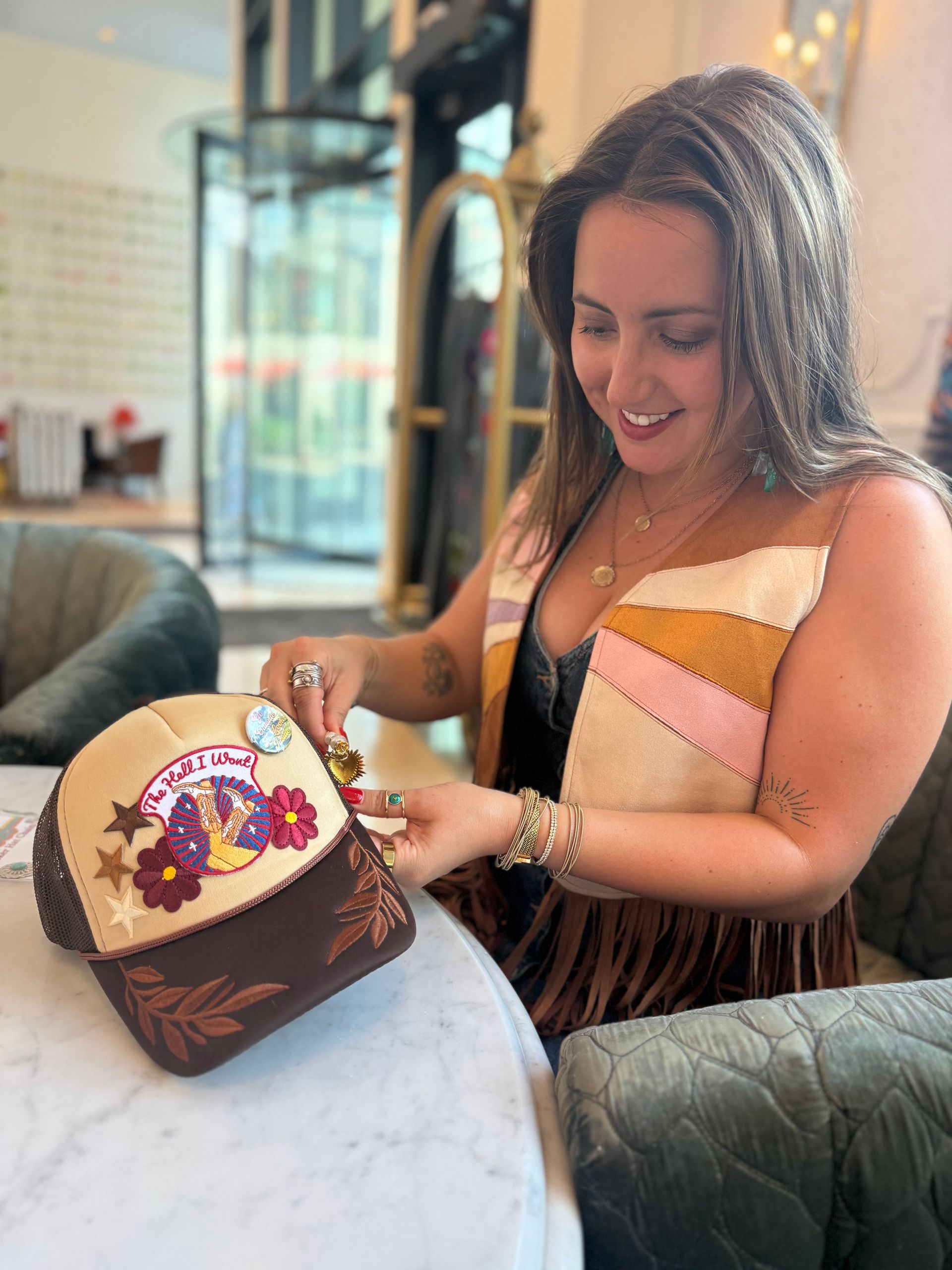Smiling woman in a fringe vest pins charms onto a beige-and-brown embroidered trucker hat at a marble cafe table