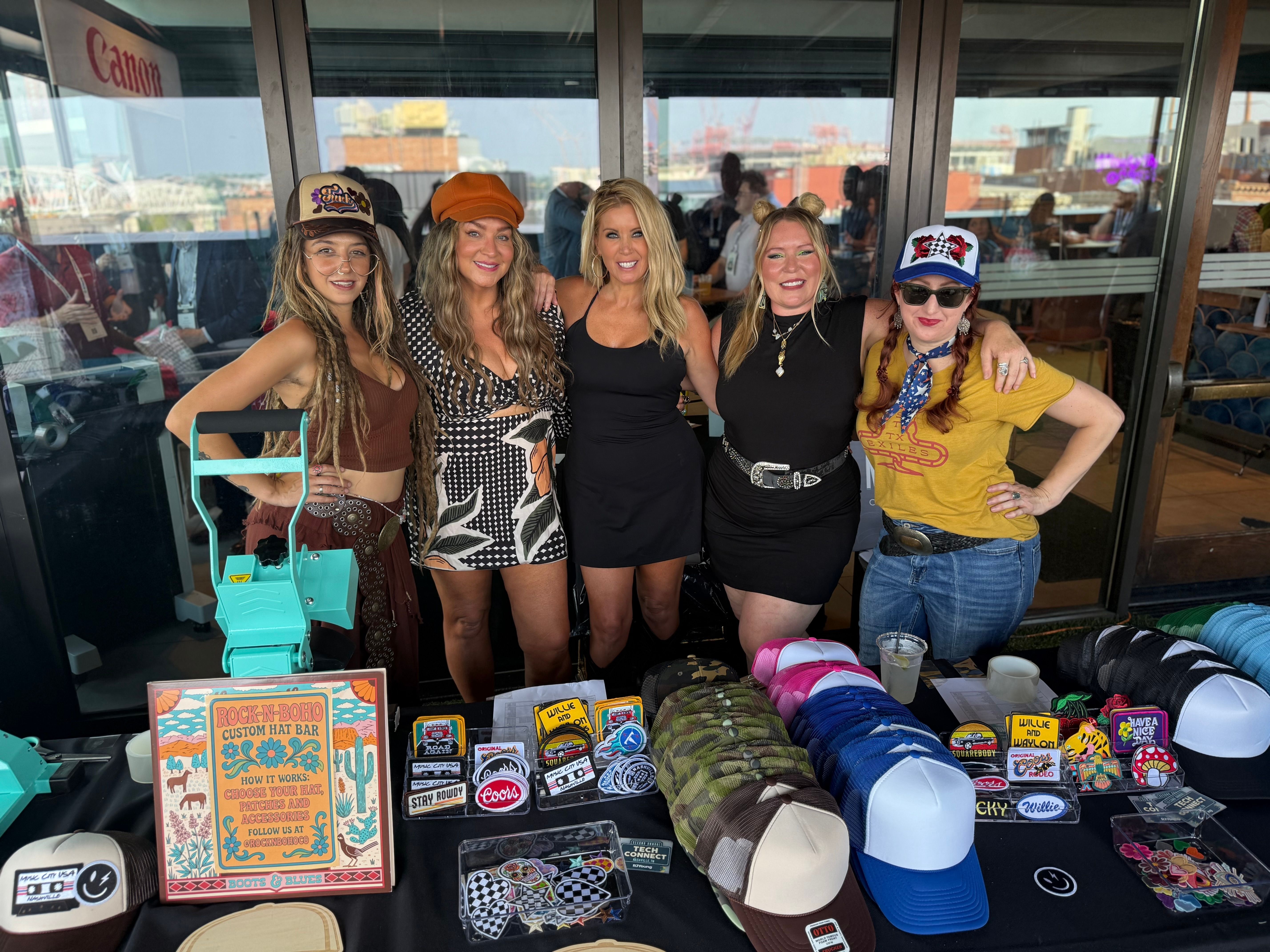 Five women smiling at a rooftop pop-up hat booth with colorful trucker caps, patches and a “custom hat bar” sign, city skyline visible through glass