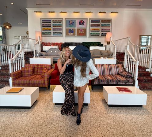 Two women laughing in a colorful boutique hotel lobby, posed in front of striped, plaid and floral sofas, white cube coffee tables, twin staircases and framed retro logo artwork.