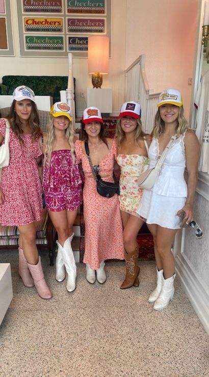 Five smiling women in colorful summer dresses, cowboy boots and trucker hats posing inside a pastel retro cafe with a striped sofa, vintage fast-food posters and a clothing rack.