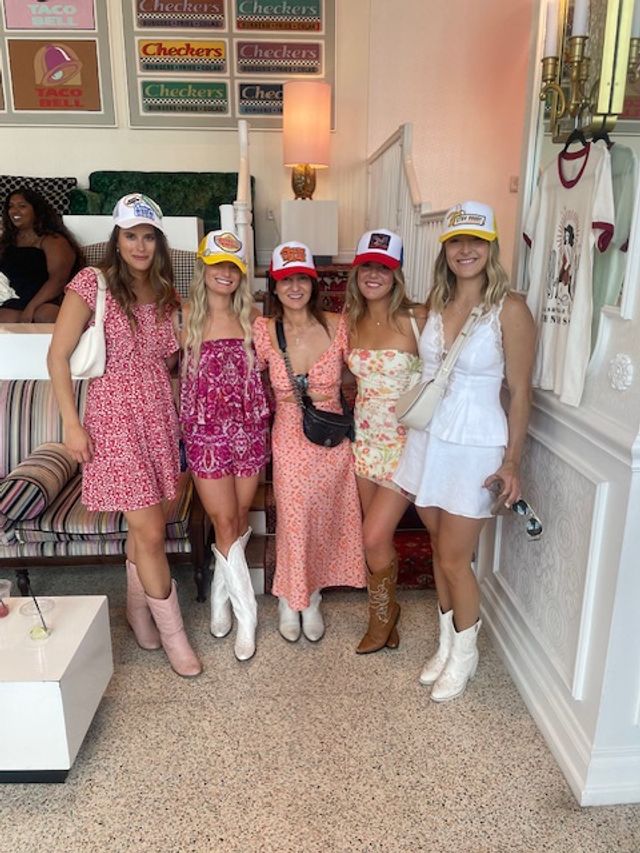 Five smiling women in colorful summer dresses, cowboy boots and trucker hats posing inside a pastel retro cafe with a striped sofa, vintage fast-food posters and a clothing rack.