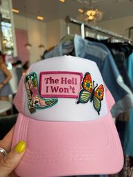 Pink trucker hat with embroidered patches — a floral cowboy boot, colorful butterfly, and a pink patch reading "The Hell I Won't," held by a hand with yellow nail polish in a boutique showcasing women's fashion accessories.