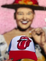 Close-up of a red, white and blue trucker hat featuring a red tongue logo, held against a blurred colorful mural portrait background for a playful street-style vibe.