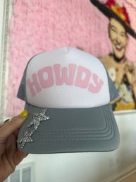 Gray-and-white trucker hat with pink "HOWDY" text and rhinestone star on the brim, held by a hand with yellow nail polish against a pink textured wall and retro portrait backdrop.