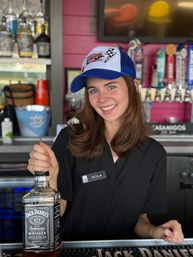Cheerful bartender in a blue trucker hat and black shirt holding a Jack Daniel's Tennessee Whiskey bottle behind a colorful bar with draft taps.